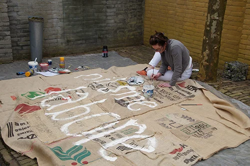hier zie je een procesfoto van het project Bunkeren. De Bunker is te zien in Arendonk, De Brulen 40. Met de hand aan elkaar gemaakte jute is de bunker helemaal bekleed. Op de jute zijn met acryl,spreuken in het Latijn geschilderd die relateren aan de landbouw. De Nederlandse betekenis is eveneens te zien, maar daar moet je even goed voor zoeken. Het is een project van Academie Arendonk en het heet Kunst in het veld, waaraan een mooie fietsroutes zijn gekoppeld