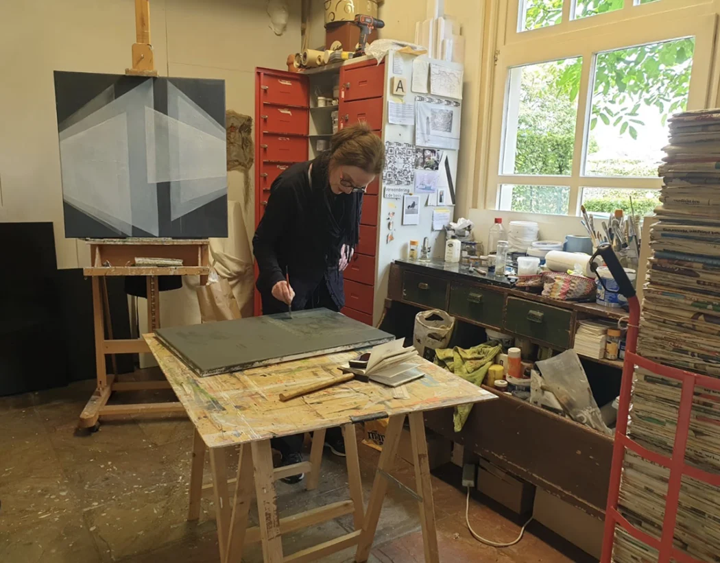 Hier ben ik aan het werk in mijn atelier. Op de achtergrond is mijn logo in wording zichtbaar. ik ben aan het werk met pigmenten waarmee ik olieverf heb gemaakt. Op de voorgrond is een toren van kranten zichtbaar, die anderhalve meeter hoog is.